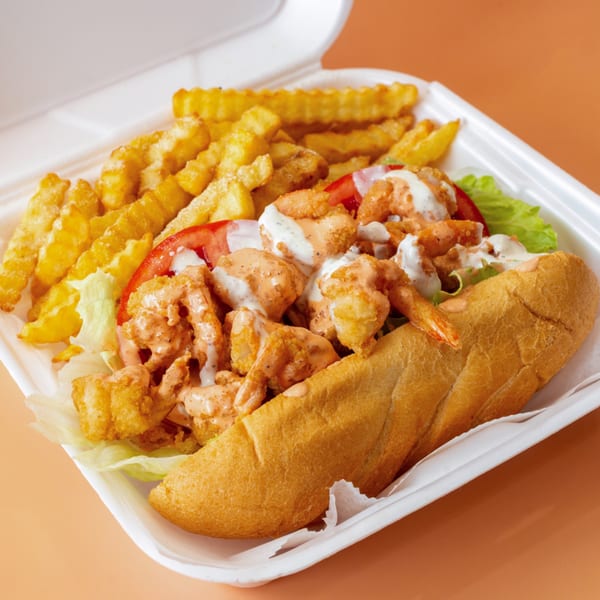 Shrimp Po'Boy sandwich with lettuce, tomato, and sauce, served with crinkle-cut fries in a white container.